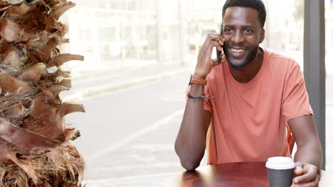 Un-Joven-Afroamericano-Habla-Por-Teléfono-En-Una-Cafetería-Al-Aire-Libre-Con-Espacio-Para-Copiar