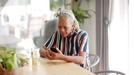 Afroamerikanische-Seniorin-Sitzt-Am-Tisch-Und-Schaut-Aufs-Smartphone