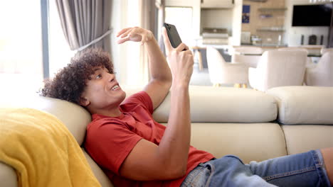 A-young-biracial-man-is-lounging-on-a-sofa-at-home,-interacting-with-his-smartphone