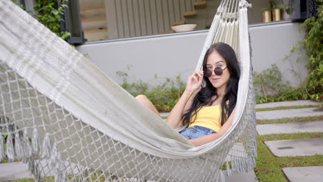 A-young-biracial-woman-lounges-in-a-hammock-in-the-backyard-at-home,-adjusting-her-sunglasses