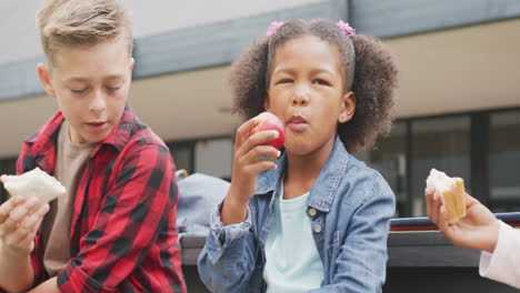 Video-of-three-diverse-schoolchildren-eating-packed-lunches,-talking-in-schoolyard