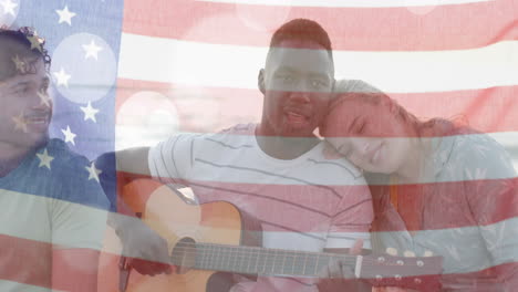 Animation-Der-Flagge-Der-USA-über-Fröhlichen,-Unterschiedlichen-Freunden,-Die-Im-Sommer-Am-Strand-Gitarre-Spielen