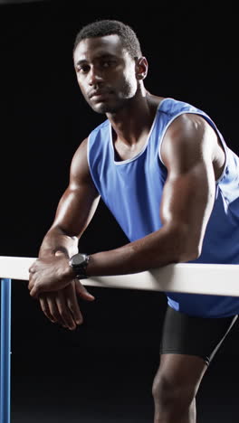 Vertical-video:-African-American-male-athlete-leaning-on-hurdle,-black-background