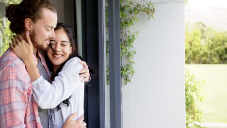 Biracial-woman-and-Caucasian-man-laughing-by-window,-embracing,-copy-space