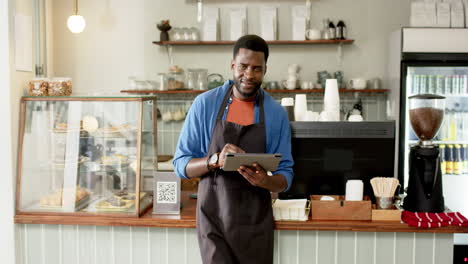 Un-Joven-Barista-Afroamericano-Se-Encuentra-Detrás-Del-Mostrador-De-Una-Cafetería,-Sosteniendo-Una-Tableta