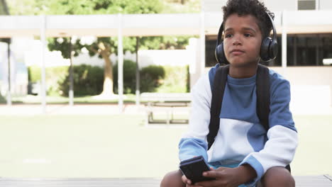 In-a-school-courtyard,-a-young-African-American-student-sits-with-headphones,-with-copy-space