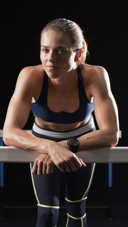 Vertical-video:-Caucasian-female-athlete-leaning-on-hurdle,-black-background