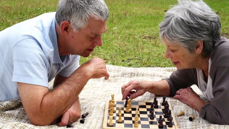 Älteres-Paar-Entspannt-Im-Park,-Liegt-Auf-Einer-Decke-Und-Spielt-Schach