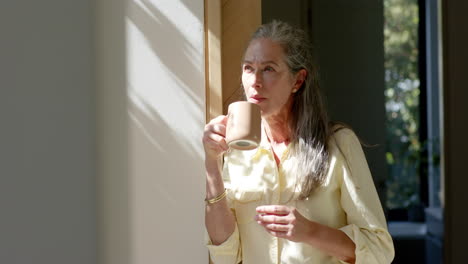 Caucasian-woman-standing-by-window,-drinking-from-cup