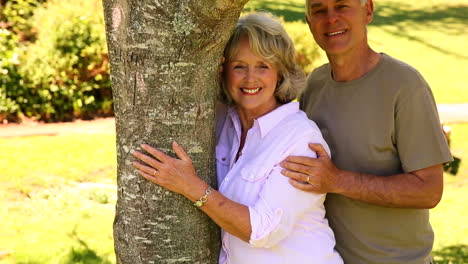 Retired-couple-leaning-against-tree-smiling-at-camera