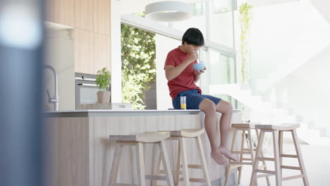 Asian-male-college-student-sitting-on-kitchen-counter,-drinking-from-bowl