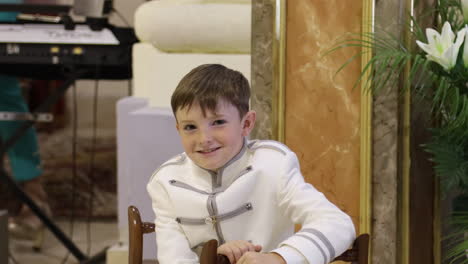 Un-Niño-Con-Un-Traje-Ceremonial-Blanco-Sentado-Y-Sonriendo-Durante-La-Celebración-De-La-Comunión.