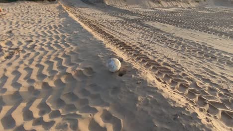 an-old-scruffy-soccer-ball-rolling-along-beach-sand-with-tire-imprints-in-the-sand-on-a-sunny-day