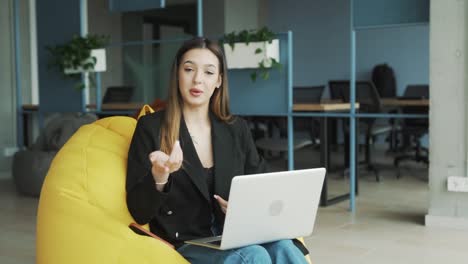 pretty-young-woman-in-business-attire-sits-on-a-pouf-with-a-laptop,-looking-at-the-camera-and-talking-against-the-background-of-a-stylish-modern-office