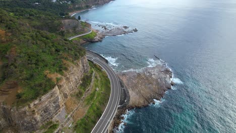 4K-Drohnenvideo-Mit-Schwenk-Nach-Unten,-Um-Die-Unglaubliche-Aussicht-Auf-Die-Sea-Cliff-Road-Zu-Zeigen.-Der-Fokus-Liegt-Dabei-Auf-Der-Straße,-Die-Sich-Neben-Dem-Atemberaubenden-Blauen-Ozean-Um-Die-Wunderschöne-Australische-Landschaft-Windet.