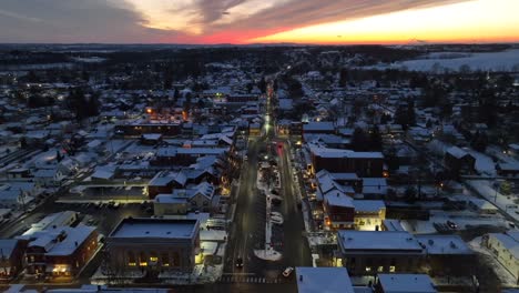 Vista-Aérea-De-Una-Ciudad-Cubierta-De-Nieve-Al-Atardecer,-Destacando-Las-Brillantes-Luces-De-La-Calle-Y-Una-Vívida-Puesta-De-Sol.