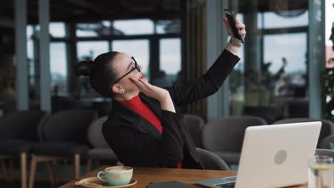 Eine-Schöne-Frau-In-Business-Kleidung-Führt-Einen-Videoanruf-Auf-Ihrem-Smartphone-Und-Lächelt,-Während-Sie-An-Ihrem-Laptop-Sitzt