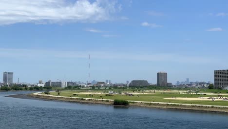 Blick-Auf-Die-Skyline-Einer-Stadt-über-Einen-Fluss-Mit-Einer-Rasenfläche-Im-Vordergrund-An-Einem-Sonnigen-Tag