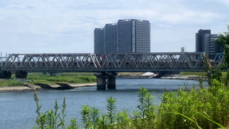 Train-crosses-a-steel-bridge-over-a-calm-river-on-a-sunny-day-with-tall-buildings-in-the-background