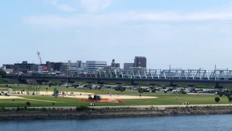 Gente-Disfrutando-De-Un-Día-Soleado-En-Un-Parque-Junto-Al-Río-Con-Edificios-De-La-Ciudad-Y-Un-Puente-Al-Fondo