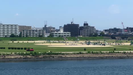 Urban-park-by-the-river-with-people-enjoying-sunny-day-and-cityscape-in-the-background