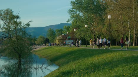 Gente-Paseando-Por-Un-Pintoresco-Sendero-Junto-Al-Lago-Con-Montañas-Al-Fondo-En-El-Lago-Jarun,-Zagreb,-Croacia