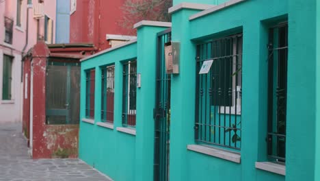 Brightly-colored-turquoise-building-with-iron-barred-windows-and-a-gate-in-a-narrow-alleyway-on-Burano-Island,-Venice,-Italy