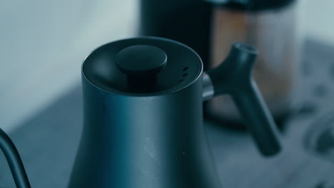 Close-up-shot-of-Hand-opening-black-Barista-Kettle-Lid-with-Coffee-Grinder-in-the-background