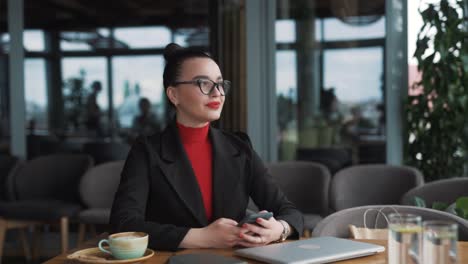 A-beautiful-young-woman-in-business-attire-sits-in-a-stylish-restaurant,-playing-a-game-or-texting-on-her-smartphone-and-smiling-cheerfully