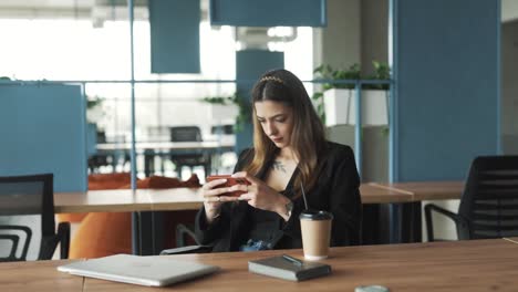 pretty-young-woman-in-business-attire-sits-in-a-stylish-modern-coworking-office,-cheerfully-and-attentively-playing-a-game-on-her-smartphone-and-rejoicing-in-her-victory