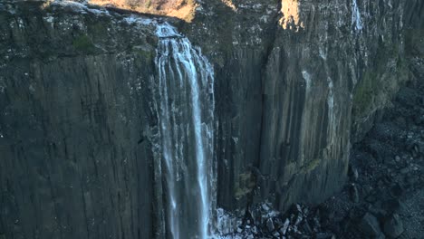 Waterfall-half-speed-slow-motion-orbit-in-shadow-falling-down-sheer-cliff-to-frosted-rocky-beach-below-in-winter-at-Kilt-Rock-Waterfall,-Isle-of-Skye,-Western-Highlands,-Scotland,-UK