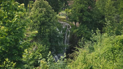 Exuberante-Vegetación-Con-Una-Cascada-Al-Fondo-Y-Gente-Disfrutando-De-Las-Vistas-Panorámicas-En-Rastoke,-Croacia