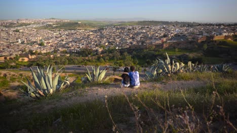 Adventurous-couple-of-man-and-woman-sit-on-dry-hillside-overlooking-Fez-Morocco
