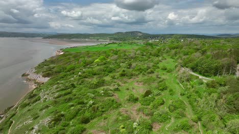 Flying-over-rugged-green-bush-dotted-coastline-towards-patchwork-fields-with-cloudy-horizon-on-bright-day-in-Spring-at-Jenny-Brown's-Point,-Silverdale,-Lancashire,-England,-UK