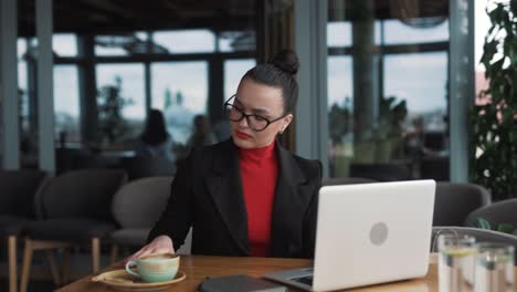 Schöne-Frau-In-Business-Kleidung-Wird-Durch-Eine-Smartphone-Benachrichtigung-Von-Ihrer-Arbeit-In-Einem-Restaurant-Mit-Einem-Laptop-Abgelenkt