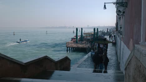 People-walking-along-a-waterfront-promenade-in-Venice,-with-boats-docked-by-the-pier-and-a-view-of-the-calm-sea