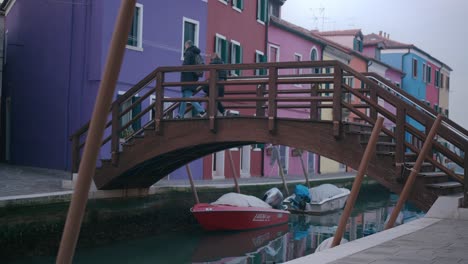 Bunte-Häuser-Säumen-Einen-Kanal-In-Burano,-Mit-Booten,-Die-Unter-Einer-Malerischen-Holzbrücke-Vertäut-Sind