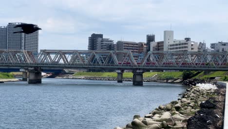 Ein-Zug-überquert-Eine-Brücke-über-Einen-Fluss-In-Einer-Städtischen-Stadtlandschaft-Unter-Einem-Klaren-Himmel