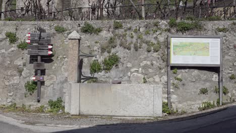 A-water-fountain,-wooden-hiking-trail-signs-and-a-map-of-the-area-in-the-village-of-Enticlar---Niclara-by-Kurtatsch---Cortaccia,-South-Tyrol,-Italy