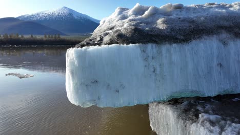 Ein-Großer-Eisblock-Hängt-über-Einem-Braunen-Fluss,-Im-Hintergrund-Ist-Ein-Schneebedeckter-Berg-Zu-Sehen,-Aufgenommen-An-Einem-Klaren-Wintertag