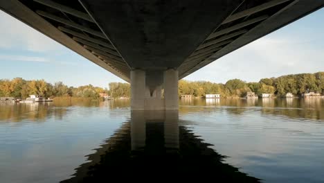 Drone-footage-of-a-calm-river-in-Europe-shows-the-underside-of-a-bridge,-with-houseboats-and-autumn-trees-lining-the-riverbanks