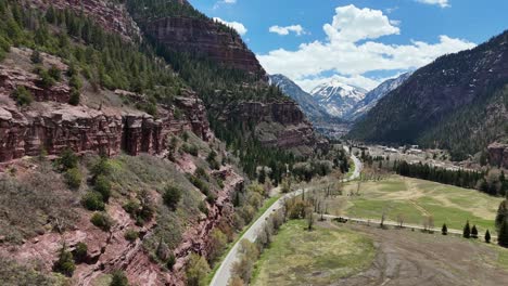 Aerial-View-of-Highway-Winding-Through-the-Mountains,-rocky-jagged-cliffs-on-one-side-of-the-road-with-pine-trees,-partial-blue-sky-with-clouds-in-background,-Ouray-Colorado