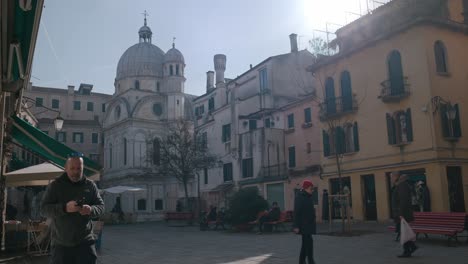 Tranquila-Plaza-De-Venecia-Con-La-Impresionante-Iglesia-De-Santa-María-Dei-Miracoli-Al-Fondo,-Gente-Paseando-Y-Sentada-En-Bancos,-El-Ambiente-Sereno-De-La-Ciudad