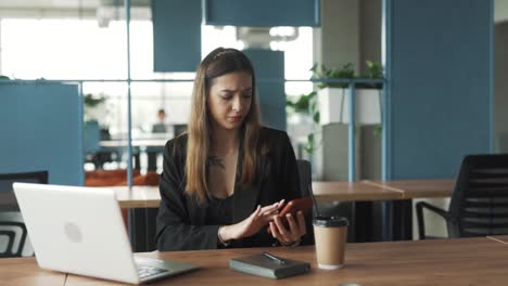 pretty-young-woman-in-business-attire-sits-in-a-stylish-modern-coworking-office,-annoyed-by-messages-on-her-smartphone-distracting-her-from-work-on-her-laptop