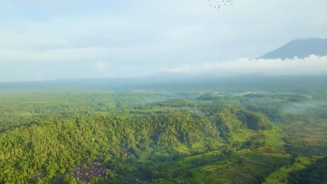 Cloudy-volcano-Mt-Agung-during-sunrise,-smokey-village-and-fields-in-the-valley,-paning-to-right-with-sunrise-rays-and-bird-flock-flying-trough