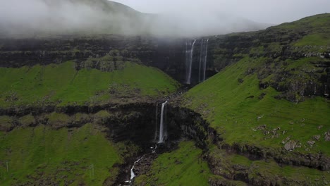 Fossá-Waterfall-with-power-line-over-river-in-cloudy-Faroe-Islands,-aerial-pull-away