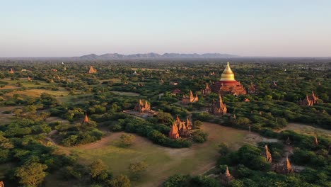 Flying-over-the-beautiful-lush,-green-landscape-of-the-countryside-of-Myanmar,-approaching-the-Dhammayazaka-Pagoda-with-many-small-temple-structures-surrounding-it---Aerial-shot