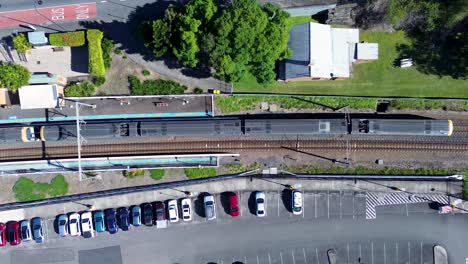Landscape-of-train-pulling-out-of-heritage-railway-station-tracks-line-surrounded-by-cars-parked-in-commuter-carpark-suburbs-town-Ourimbah-Australia-drone-aerial