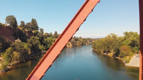 Drone-flying-through-the-Fair-Oaks-Bridge-sideways-with-a-view-of-the-American-River-surrounded-by-green-trees---California