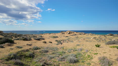 A-wide-view-of-the-sandy-terrain-at-the-Tombs-of-the-Kings-in-Pafos,-Cyprus,-with-the-sea-in-the-background-and-a-partly-cloudy-sky